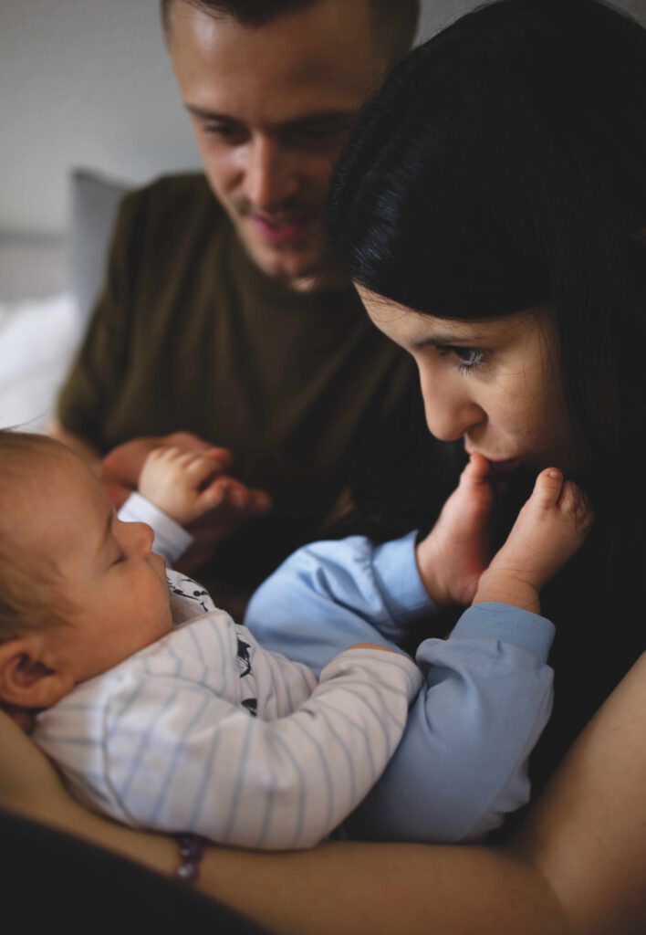 Mama hat Baby auf dem Schoß, küsst die Füße, Papa sitzte daneben und hält die Hand des Babys